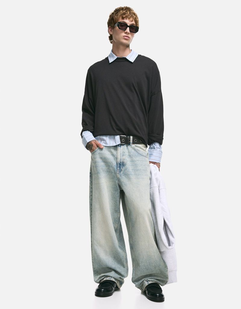 Full-length studio shot: adult male in sunglasses, black sweater over collared shirt, baggy jeans, and loafers on white background.