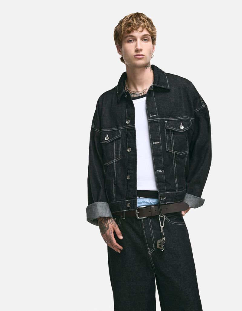 Male fashion model wears a dark denim jacket and jeans in a studio shot, showcasing contemporary style and casual wear against a white backdrop.
