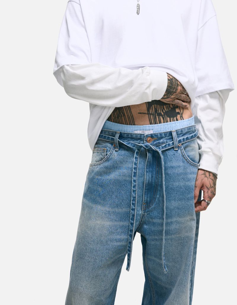 Fashionable medium studio shot of person wearing baggy blue jeans with tied belt, a long-sleeved white shirt, and a silver pendant necklace.