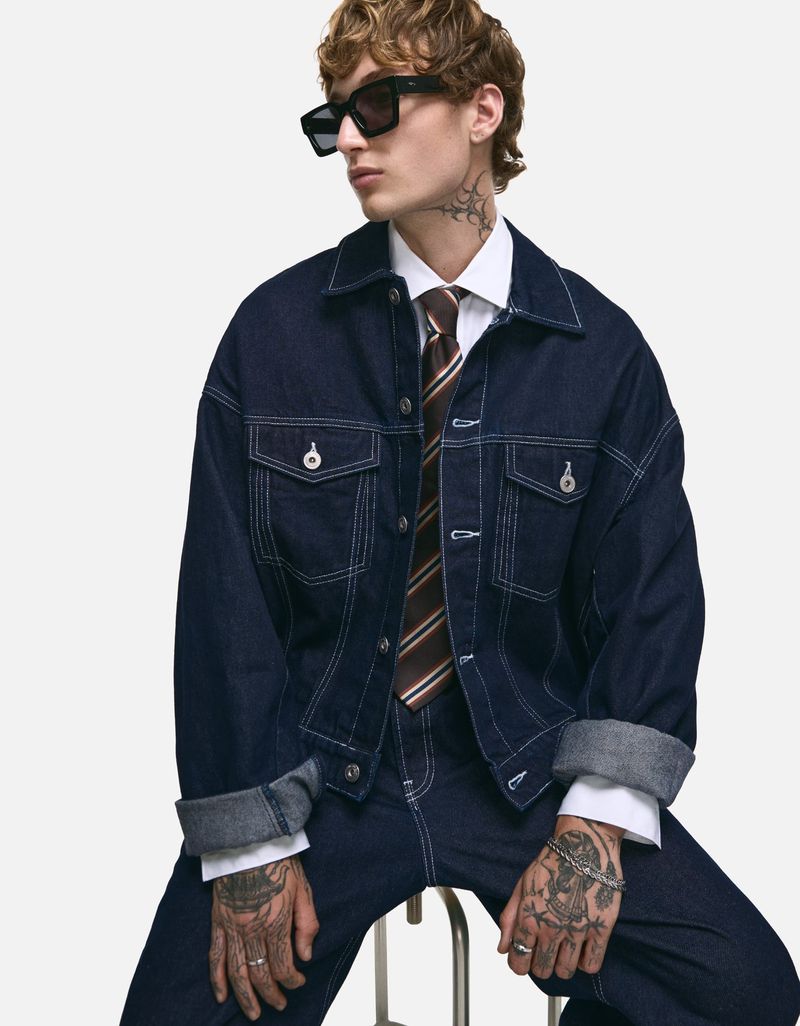 A young adult man with curly hair is sitting while wearing a denim jacket with jeans, a brown tie, and black sunglasses.
