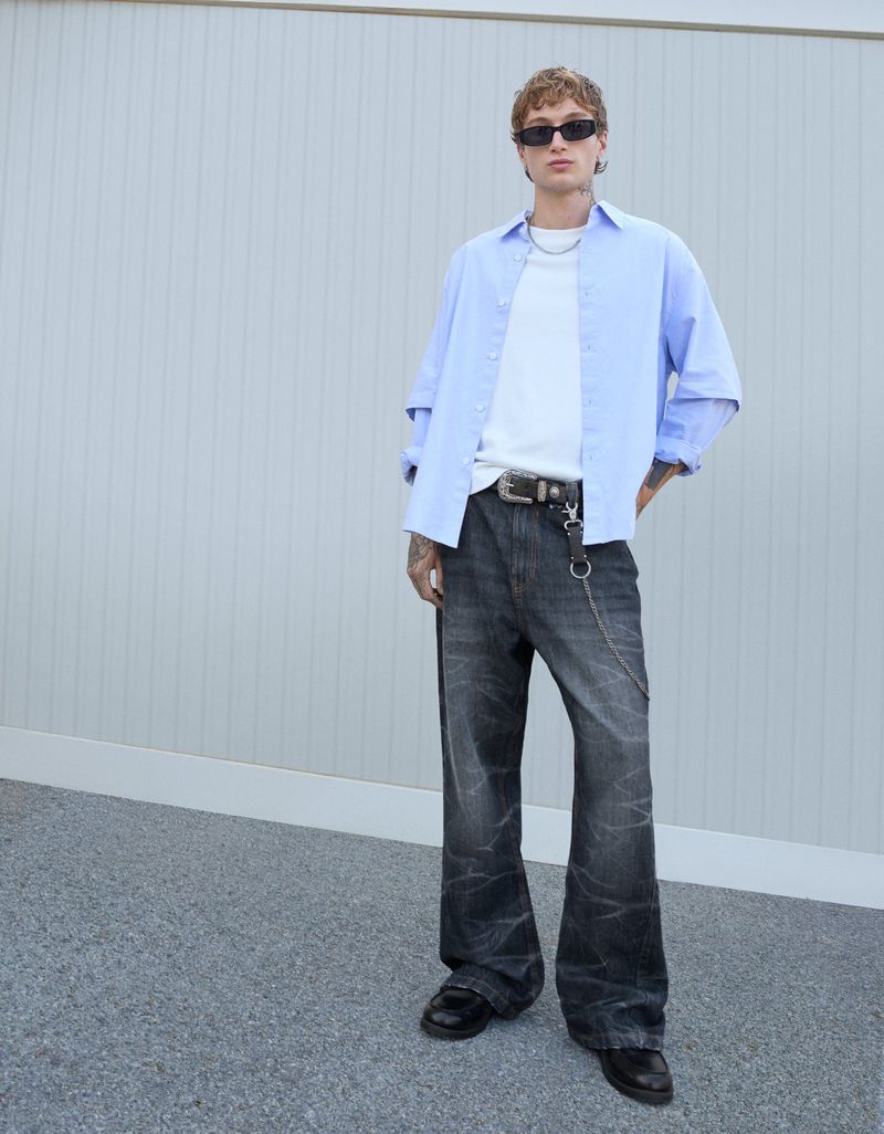 Male model in sunglasses, light blue button-down shirt, white t-shirt, wide-leg denim jeans, and black shoes poses against a plain wall.