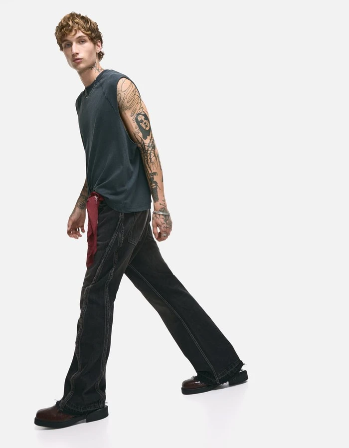 Young man with tattoos walking in a dark grey sleeveless top and black denim trousers against a white background