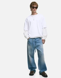 Model wearing dark sunglasses, layered white t-shirt, and baggy denim jeans against a white background.