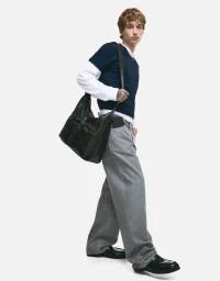 Young man wearing a layered navy short-sleeve shirt over white sleeves, grey jeans, and black loafers holding a black shoulder bag.