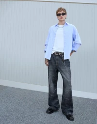 Young man wearing sunglasses, blue button-down shirt, white t-shirt, and dark patterned denim jeans standing outdoors.