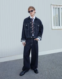 Model in dark denim jacket and matching jeans wearing sunglasses against a light architectural wall.