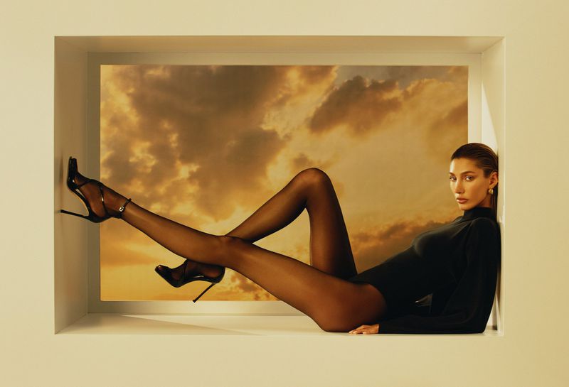 A woman in a black bodysuit and heels poses within a white window frame, set against a dramatic sunset cloudscape backdrop.