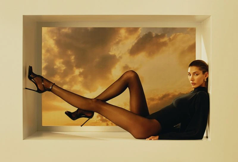 A woman in a black bodysuit and heels poses within a white window frame, set against a dramatic sunset cloudscape backdrop.