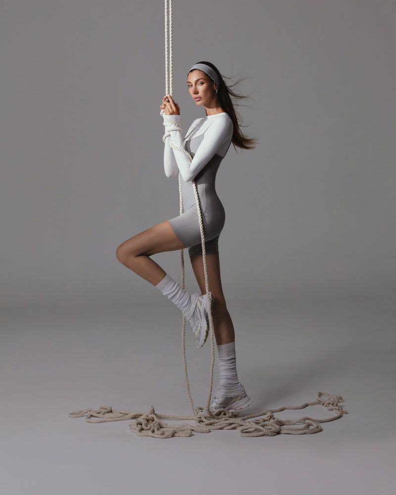 A woman in a gray bodysuit and athletic wear stands, holding a thick white rope, with a neutral gray background in a studio setting.