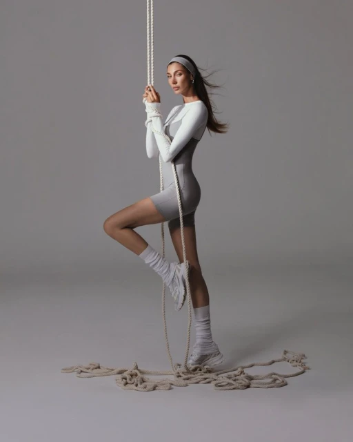 A woman in a gray bodysuit and athletic wear stands, holding a thick white rope, with a neutral gray background in a studio setting.