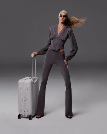 A woman in grey sportswear stands next to a grey suitcase against a studio backdrop, showcasing fashion and travel style.
