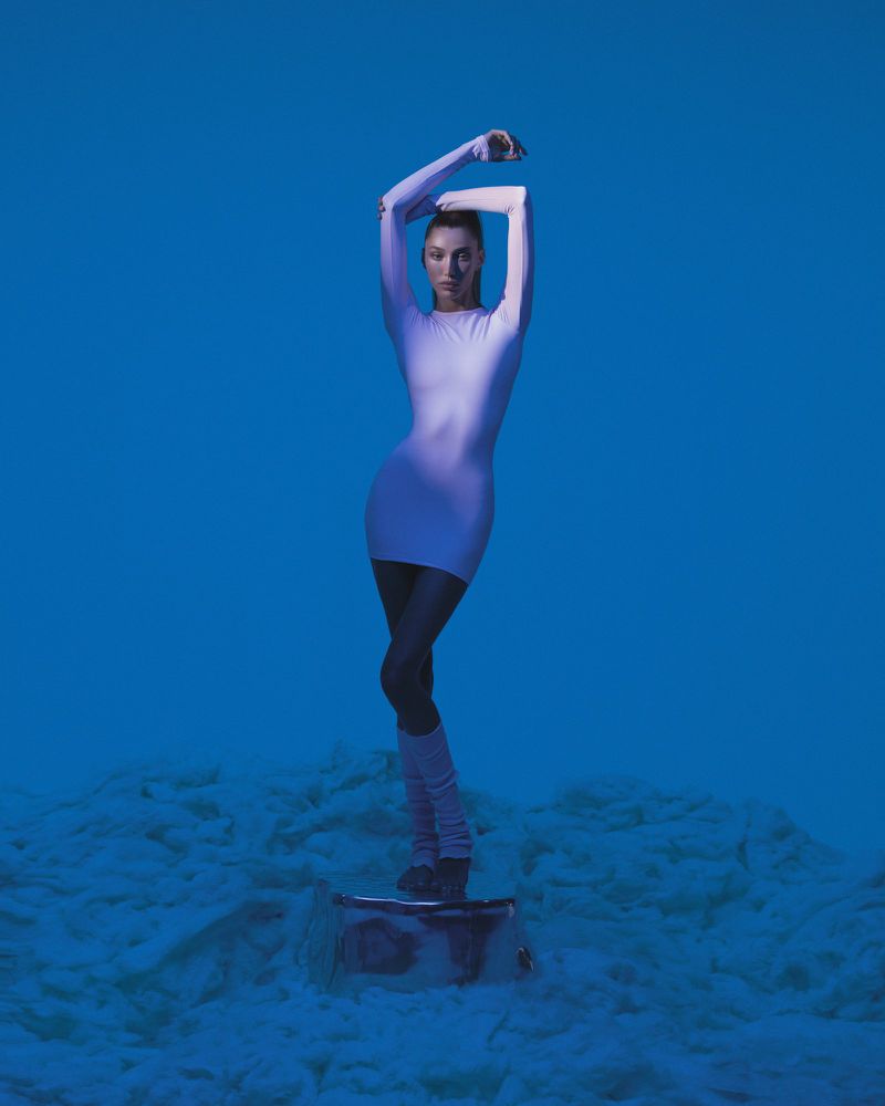 A slender woman with a high ponytail stands on a reflective surface in a short dress, leg warmers, and shoes, against a blue backdrop.