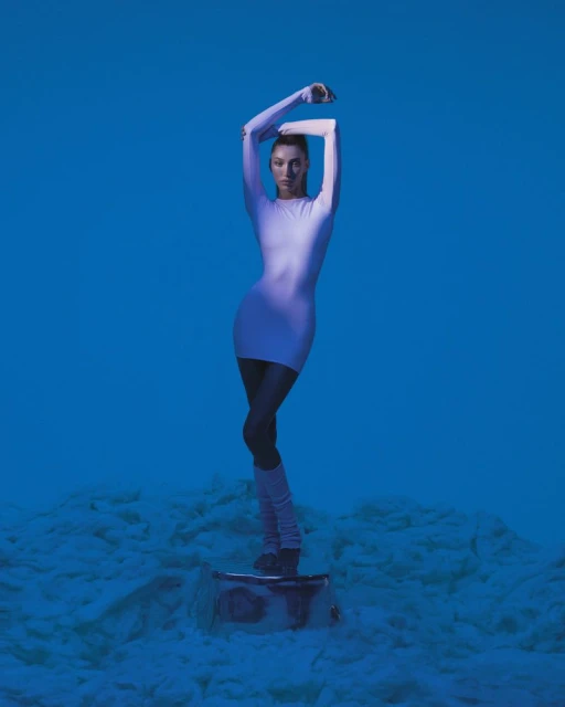 A slender woman with a high ponytail stands on a reflective surface in a short dress, leg warmers, and shoes, against a blue backdrop.