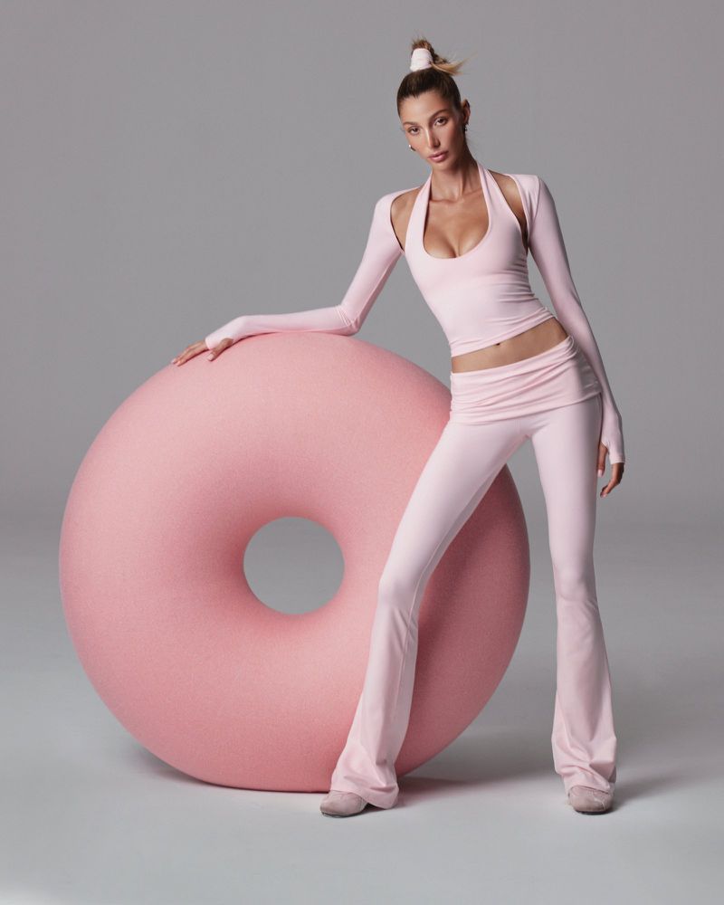 A woman in athletic wear poses beside a pink donut. She wears a matching crop top and pants, and the background is a light gray.