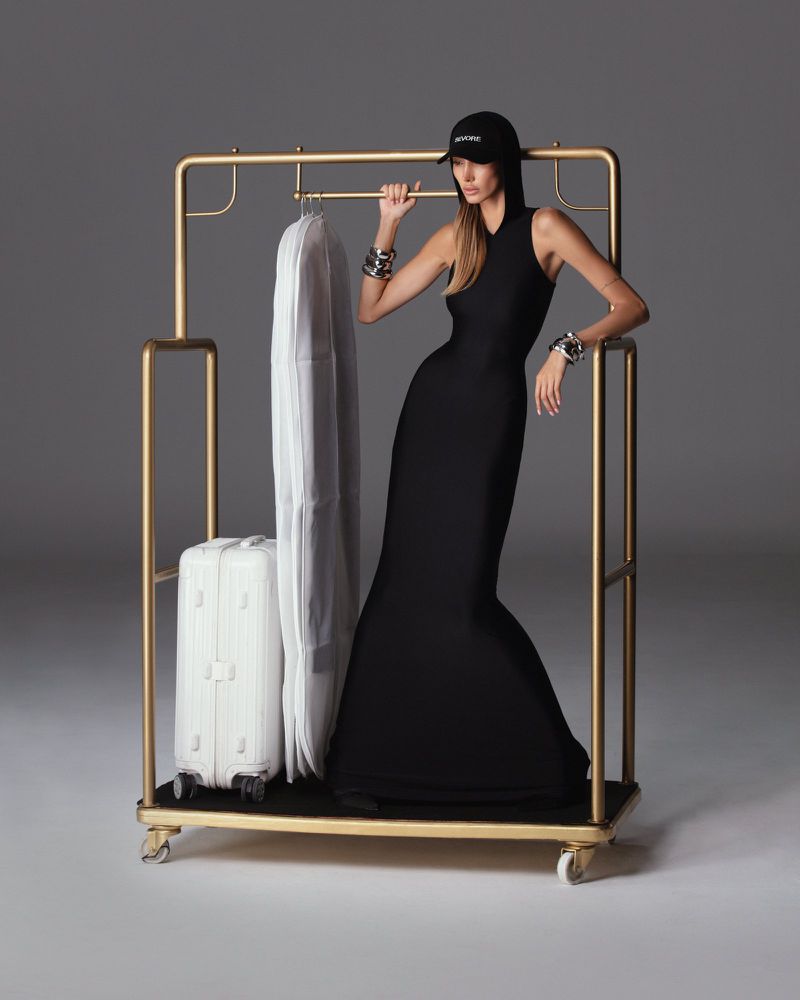 Woman in a black dress and baseball cap posing next to a gold clothing rack with a garment bag and white suitcase in a studio.