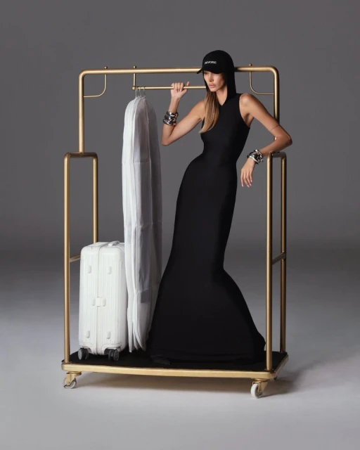 Woman in a black dress and baseball cap posing next to a gold clothing rack with a garment bag and white suitcase in a studio.