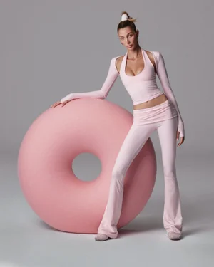 Model in pink activewear leaning on a large pink torus prop against a grey studio backdrop