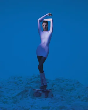 Model posing on a metallic cylinder pedestal wearing a long sleeve white dress against a monochromatic blue background
