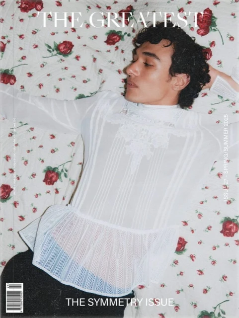 Fashion magazine cover featuring a reclining person with curly dark hair wearing a frilly, see-through white blouse on a floral bedspread.