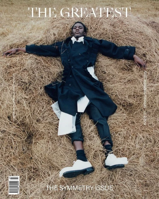 Editorial fashion shot of an African American male model in a black trench coat reclining on a pile of hay for a magazine cover.