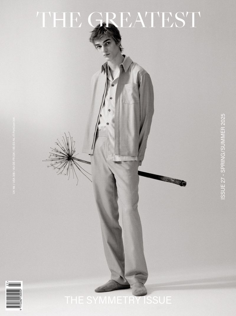A young, light-skinned male model stands in a studio with neutral lighting, wearing a linen suit, holding a unique floral branch. Spring/Summer fashion editorial.
