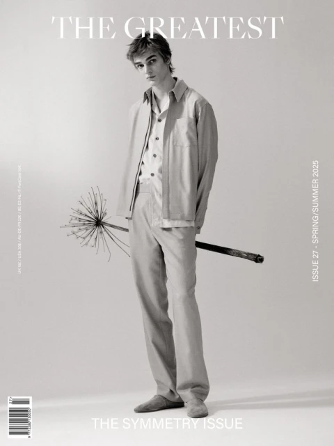 A young, light-skinned male model stands in a studio with neutral lighting, wearing a linen suit, holding a unique floral branch. Spring/Summer fashion editorial.