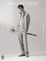 Young male model wearing an open jacket and trousers while holding a large dried flower stem against a seamless grey backdrop.