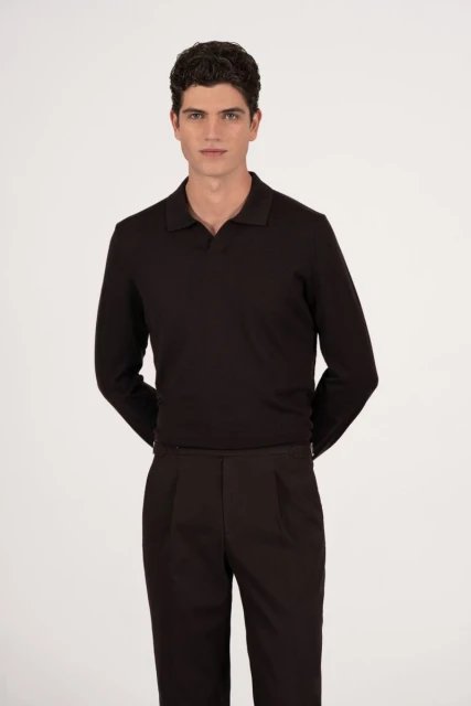 Man in black long-sleeve polo shirt and dark pants stands indoors against a white background in a waist-up studio shot.