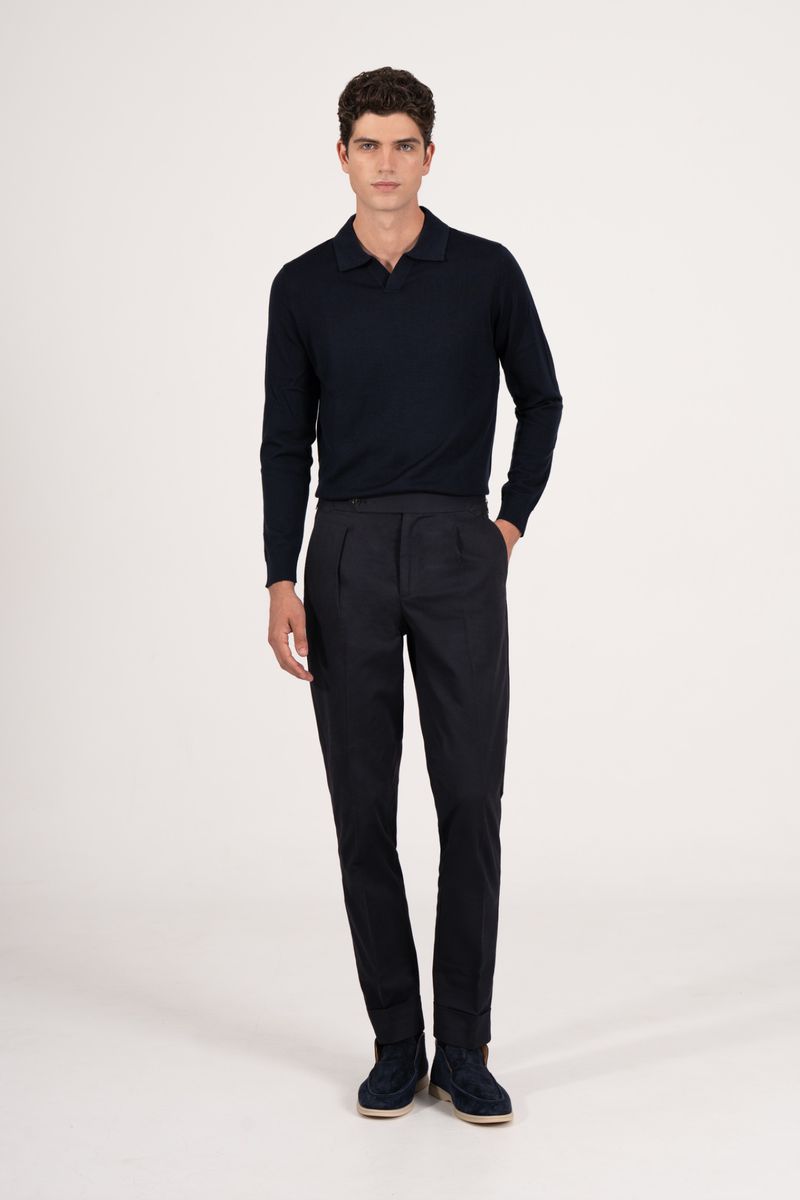 Full-length studio shot of a male model in a navy blue long-sleeved polo shirt and pleated trousers posing against a white backdrop.