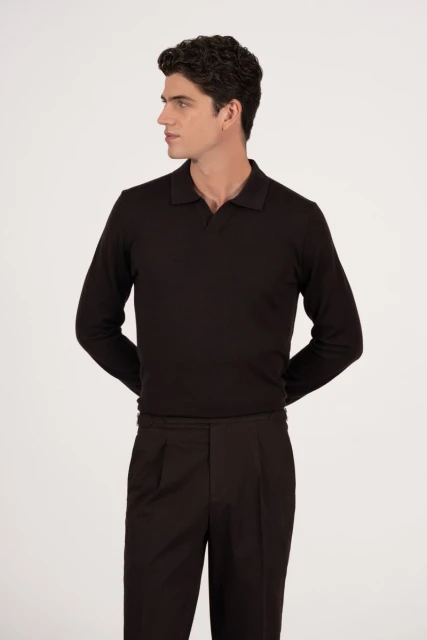 A man wearing a dark, long-sleeve polo shirt and pleated trousers poses against a neutral backdrop, looking to the side in a studio shot.