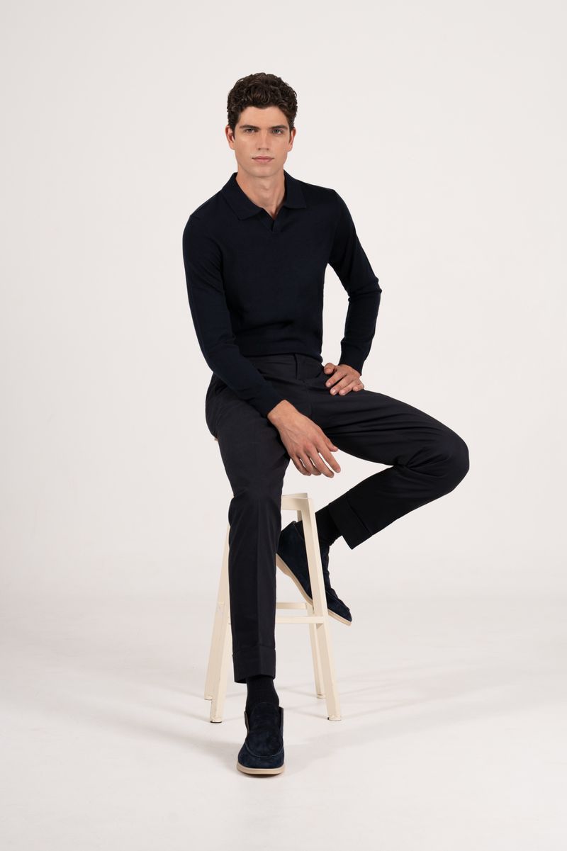 A fair-skinned adult male sits on a white stool wearing a dark navy long-sleeve polo shirt with matching pants and shoes in a studio.