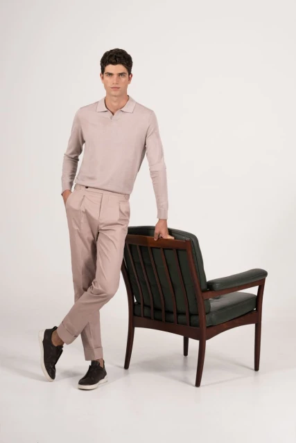 A well-dressed adult man leans against a dark-green armchair in a light studio, wearing a beige polo sweater, matching pants, and dark sneakers.
