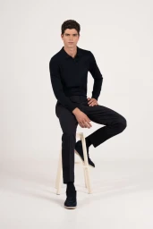 Male model seated on a white stool wearing a dark navy polo shirt and matching trousers against a white seamless background