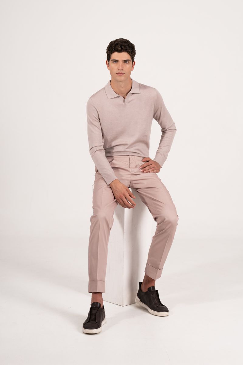 Male model seated on a white block wearing a light pink polo shirt, trousers, and dark sneakers against a white background