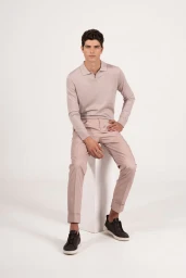 Male model seated on a white block wearing a light pink polo shirt, trousers, and dark sneakers against a white background