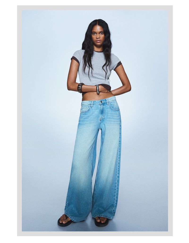A slender adult female model poses in a photo studio wearing a grey T-shirt, wide-legged light blue jeans, bracelets, and black sandals.