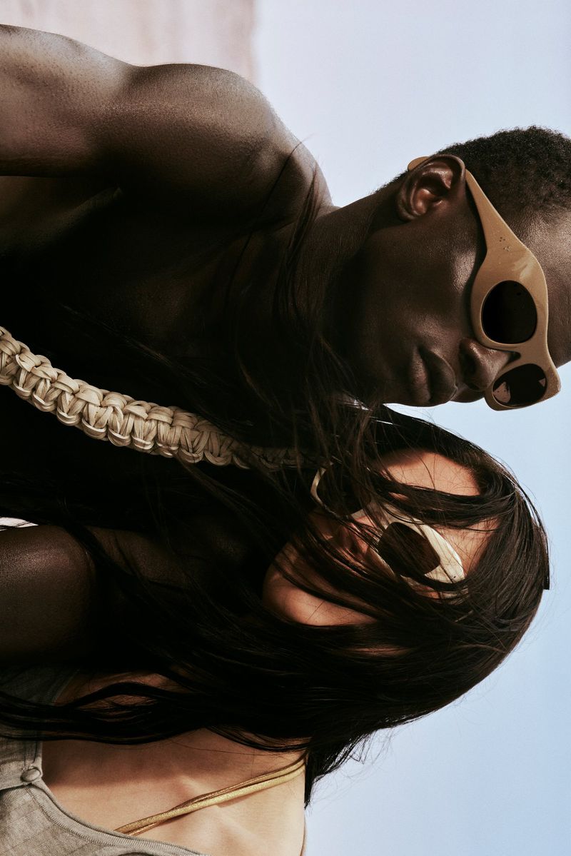 Dark-skinned adult couple wearing sunglasses, braided strap, and gray linen clothing, posing closely with a bright sky in the background.