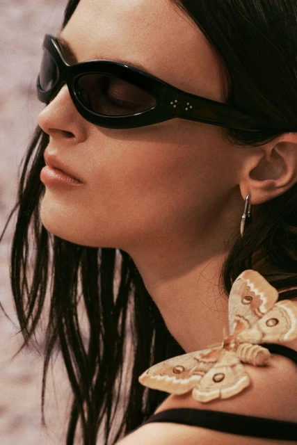 Portrait of a brunette woman wearing black sunglasses and a black top, adorned with a moth accessory, against a neutral backdrop.