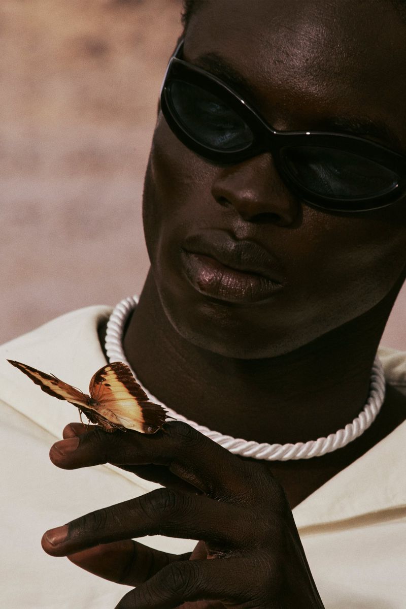Close-up of a dark-skinned man wearing sunglasses and a necklace, with a butterfly perched on his finger, against a neutral background.