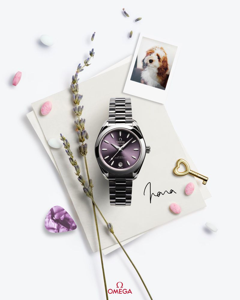 Elegant Omega wristwatch with a purple face and silver band on a white surface with lavender, candy, and a photo of a dog.
