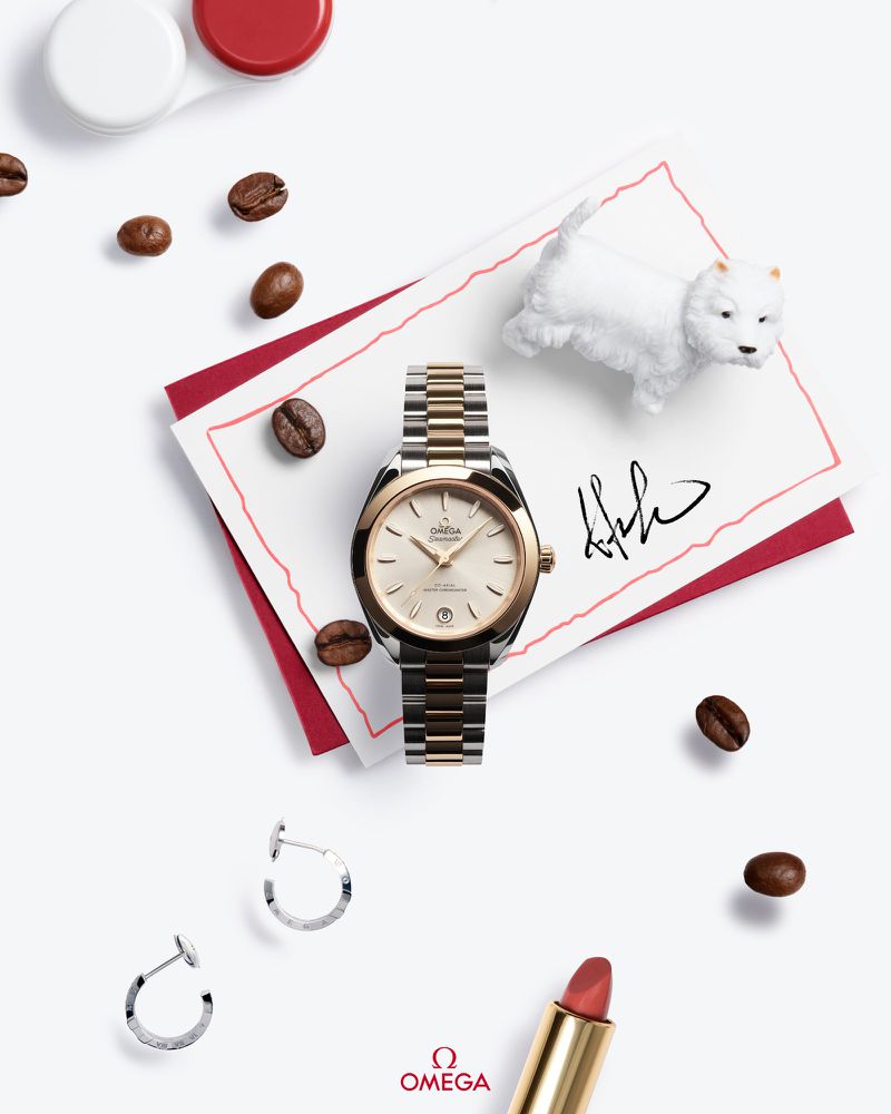 Close-up of a gold and silver Omega watch on a white surface, surrounded by coffee beans, earrings, lipstick, and a dog figurine.