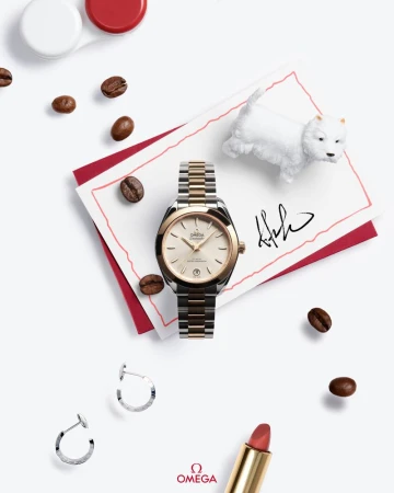 Close-up of a gold and silver Omega watch on a white surface, surrounded by coffee beans, earrings, lipstick, and a dog figurine.