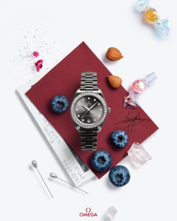 Luxury wristwatch with diamond bezel on a maroon card, surrounded by scattered blueberries, almonds, pins, and wrapped candies on white.