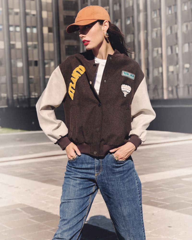 Woman in bomber jacket with a baseball cap poses outdoors in front of a modern building on a sunny day in a medium shot.
