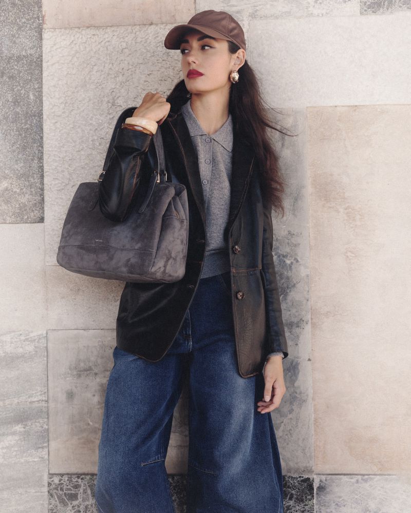 A woman in jeans, a blazer, and a baseball cap carries a large handbag while posing against a marble wall in natural light.