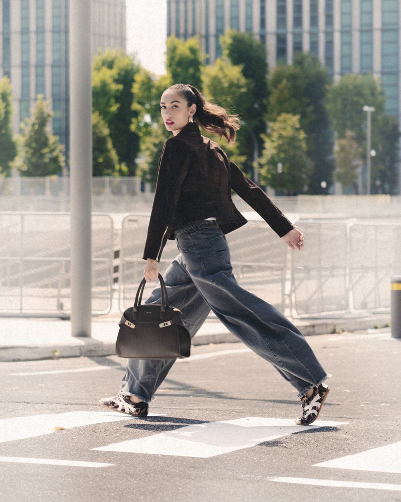 A stylish woman in a black jacket and wide-leg jeans confidently strides across a zebra crossing in an urban setting, carrying a dark handbag.