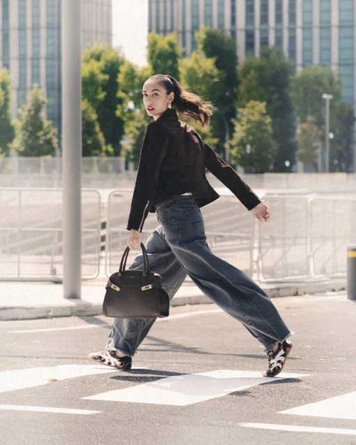 A stylish woman in a black jacket and wide-leg jeans confidently strides across a zebra crossing in an urban setting, carrying a dark handbag.