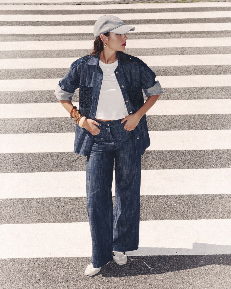 A woman in a denim outfit stands on a crosswalk, wearing a cap, blue denim jacket, and pants, with white sneakers, posing with hands in pockets.