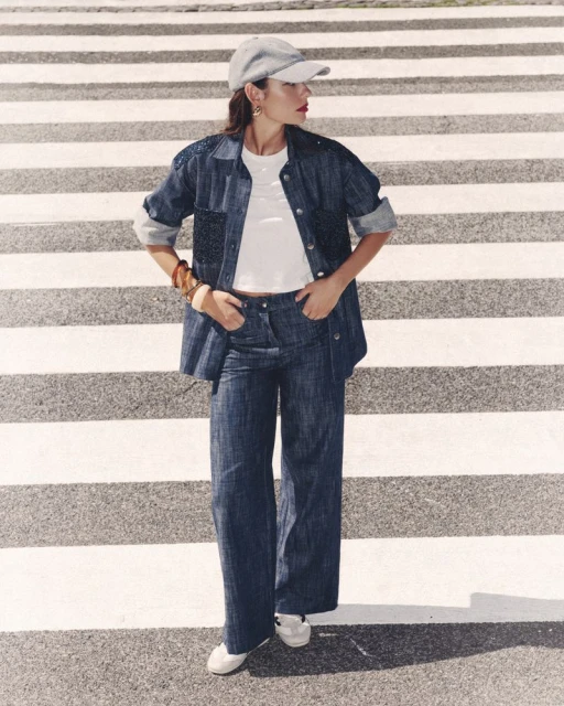 A woman in a denim outfit stands on a crosswalk, wearing a cap, blue denim jacket, and pants, with white sneakers, posing with hands in pockets.