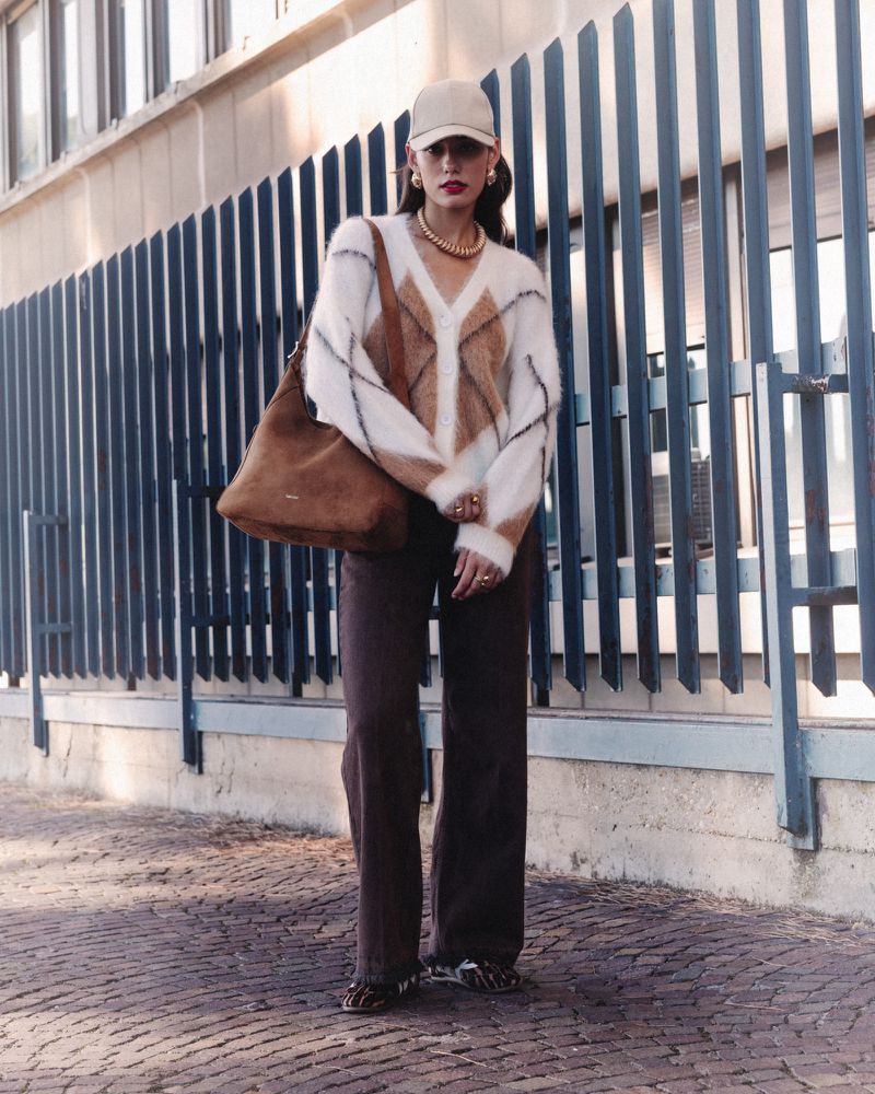 A young adult woman stands outside wearing brown pants, a white, brown and black sweater with a brown tote bag over her shoulder.